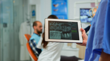 Close up dentist nurse holding tablet with digital radiography, while doctor is working with...