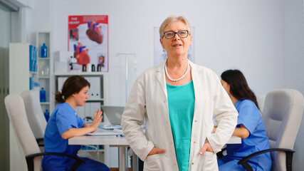 Portrait of elderly doctor standing in front of camera smiling in medical conference meeting office. Team of specialist doctors working in background talking about symptoms of disease in clinic room