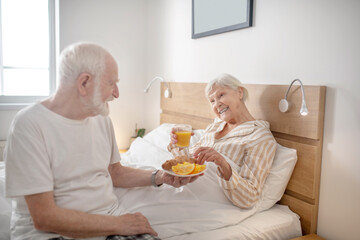 Grey-haired bearded man giving orange juice to his wife