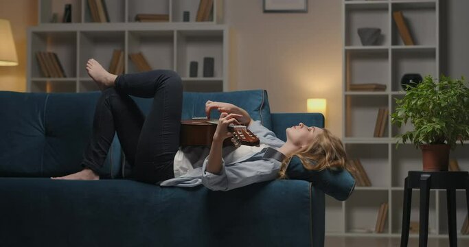 thoughtful woman is playing sad song on guitar lying on couch in cozy apartment at evening, melancholy and boring