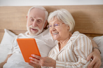Aged married couple lying in bed and having good time while reading