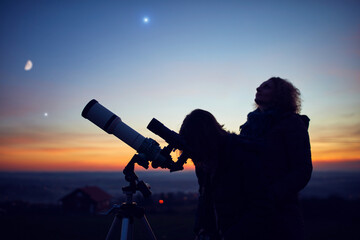Mother and daughter observing stars, planets, Moon and night sky with astronomical telescope.