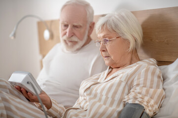 Grey-haired elderly man lying in bed and checking blood pressure with his wife