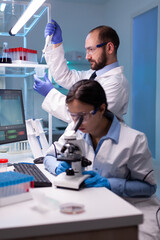 Scientist examining blood sample looking under microscope while chemist doctor using micropipette...