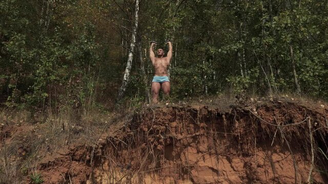 Tanned jock in sexy spandex shorts stands on the edge of a sandy cliff and stretches his arms with small dumbbells