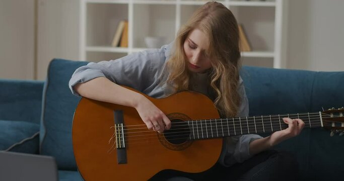 young woman is playing melody on guitar, hobby and entertainment at home at weekend, sitting on couch in living room