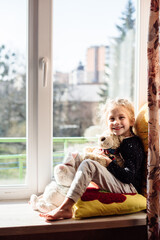 Cute little girl enjoying the sunshine while sitting at the window. The child is playing sitting on...