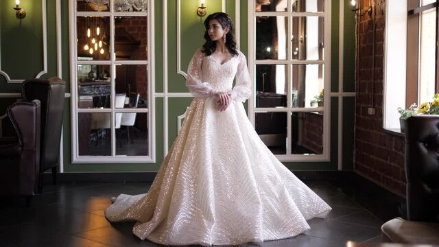 Attractive lady in elegant white decorated wedding dress with loose sleeves and trail near mirrors in restaurant slow motion