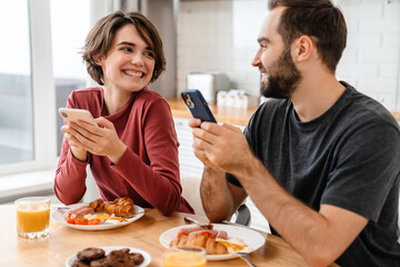 Obraz na płótnie Canvas Happy beautiful couple using cellphones while having breakfast