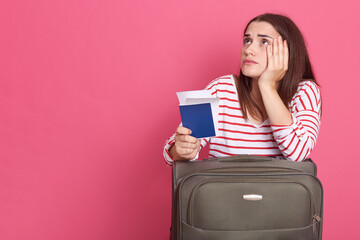 Young European traveler woman holding suitcase and documents, isolated on pink, being disappointed, looking up with sad expression, keeps hand on cheek, waits her flight, copy space for advertisement.