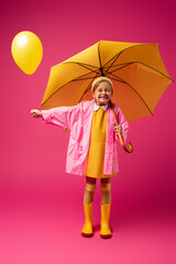 full length of excited girl in raincoat holding balloon and yellow umbrella on crimson