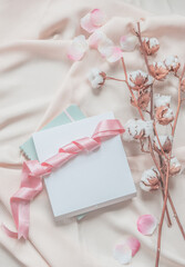 Aesthetic lifestyle with notebook, white box and pink ribbon on beige fabric background with cotton branches and pink petals. Top view. Beauty