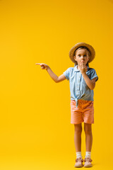 full length of shocked kid in straw hat covering mouth and pointing with finger on yellow
