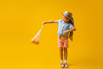 full length of excited kid in straw hat pointing with finger at reusable string bag with oranges on yellow