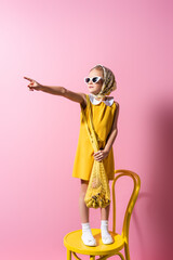 girl in headscarf and sunglasses holding reusable string bag with bananas while standing on chair and pointing with finger on pink