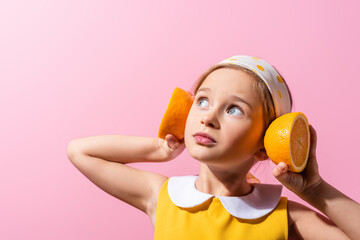 girl in headscarf holding orange halves near ears isolated on pink