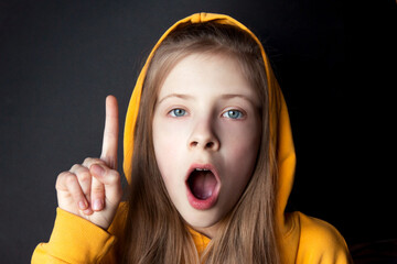 Teenager girl in an orange sweatshirt with bright emotions on her face on a black background. The finger is raised up. I have an idea. Selective focus