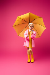 full length of happy girl in raincoat standing under yellow umbrella on crimson