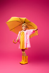 full length of girl in raincoat and rubber boots standing under yellow umbrella on crimson