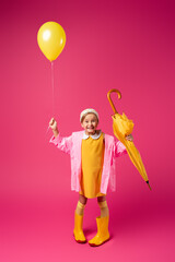 full length of happy girl in raincoat and rain boots holding balloon and yellow umbrella on crimson