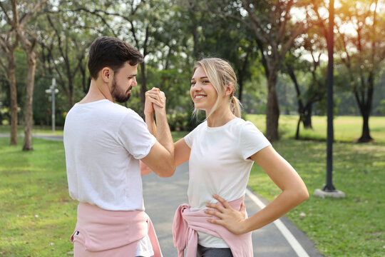 image of young caucasian couple in white and pink sport dress with smiling and arm wrestling challenge in public park outdoor with green tree in nature