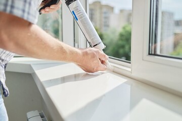 A worker with a construction syringe fills seam between sill and window with silicone sealant