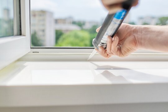 A worker with a construction syringe fills seam between sill and window with silicone sealant