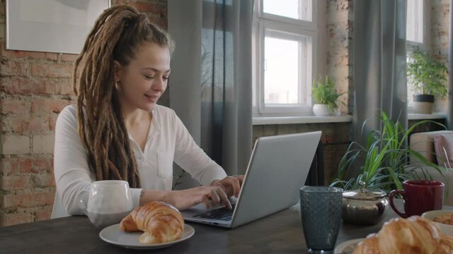 Panning slow-motion medium shot of pretty young female freelancer sitting at table, working on laptop during breakfast