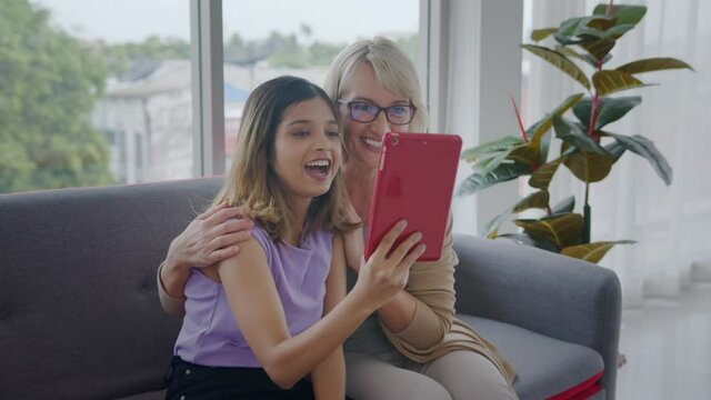 Medium shot, slow motion - Adopted Asian teen daughter and her Caucasian stepmother enjoying a video call through tablet with family members. Modern family lifestyle with internet technology.