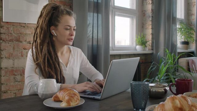 Panning slow-motion medium shot of young caucasian woman with dreadlocks sitting at kitchen table in wireless headphones, having video call via laptop during breakfast