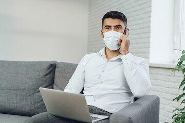 Handsome man businessman in medical mask using his laptop while talking on his cellphone
