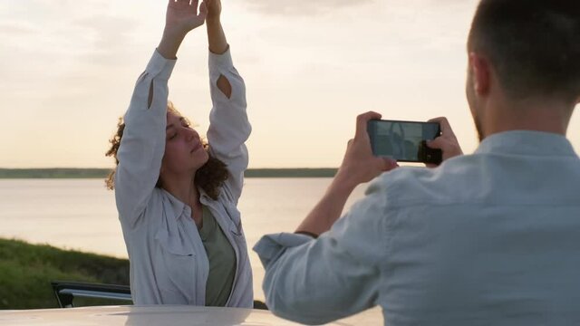 Young boyfriend taking photos of attractive girlfriend with long curly hair standing against beautiful lake