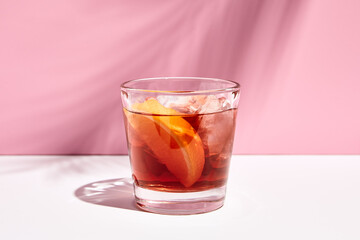 Fototapeta na wymiar Negroni cocktail over pink background. Drink in rox glass in daylight with palm leaf hard shadow. Summer, tropical, fresh drink concept