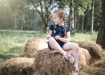 Naklejka na ściany i meble Beautiful young girl exploring nature with plaited pig tails hair on summer day sunshine sitting on bails of hay and straw in tall trees woodland forest and long grass sun rays shining in