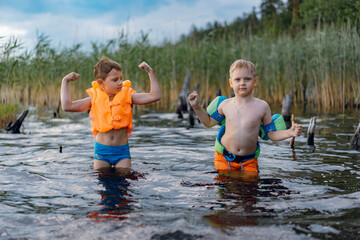 Fototapeta premium portrait of two cute little boys standing at lake shore in the forest. one boy wearing life vest, another wearing armbands. Reeds grow in water and some snags come out of pond. 