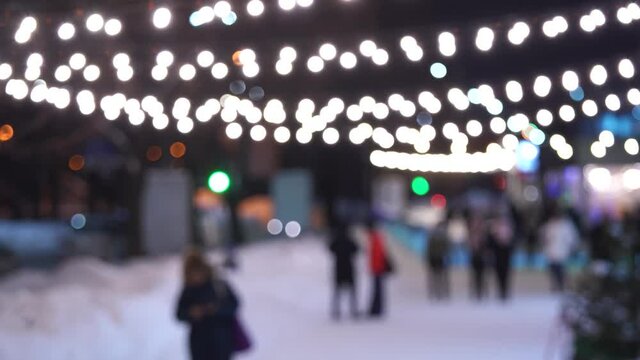 Blurry defocused winter city landscape. Silhouttes of unrecognizable people walk on snowy cold sidewalk. Bokeh of glowing holiday Christmas garlands hanging overhead. Merry winter season concept
