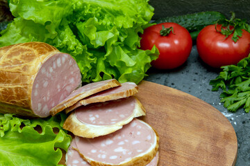 Large thick sausage on a wooden board, garnished with green salad and red tomatoes. Large sausage sliced ​​on a cutting board. Close-up.