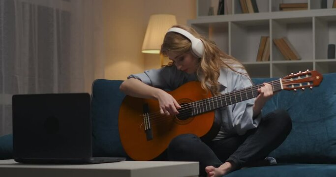 student girl is playing guitar at evening at home, listening to music by headphones and viewing notes in laptop