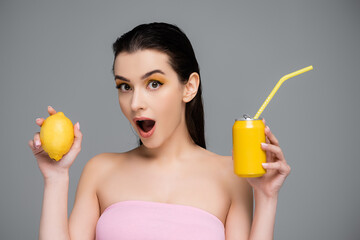 amazed young woman holding yellow can with soft drink and lemon isolated on grey