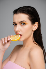 surprised young woman with bare shoulders biting sliced lemon isolated on grey