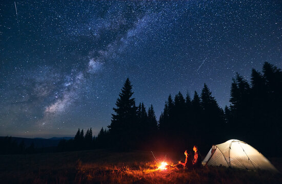 Young couple hikers sitting near bright burning bonfire and illuminated tourist tent, enjoying camping night together under dark sky full of shiny stars and bright Milky Way, warm summer evening.