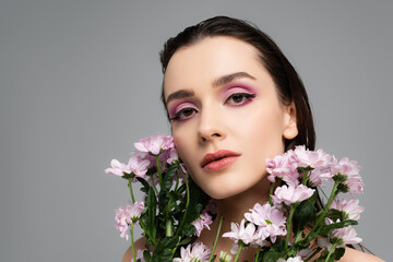 young woman with pink eye shadows looking at camera near flowers isolated on grey