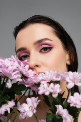 young woman with pink eye shadows looking at camera through flowers isolated on grey
