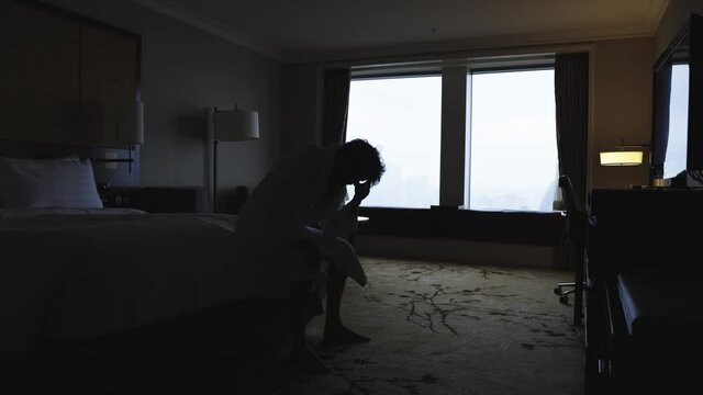 Unhappy man standing in window of hotel room, looking down to street and thinking about committing suicide. Male figure in bathrobe in backlight. Despair, mental health and depression concept.