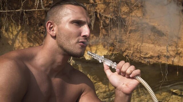 Brutal shirtless man smokes a hookah on the beach near a sandy cliff exhaling clouds of smoke