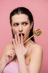shocked young woman covering mouth and holding lollipop isolated on pink