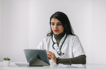 Indian female doctor in a white coat with a stethoscope conducts an online consultation in her office. Female doctor communicates with the viewer directly into the camera