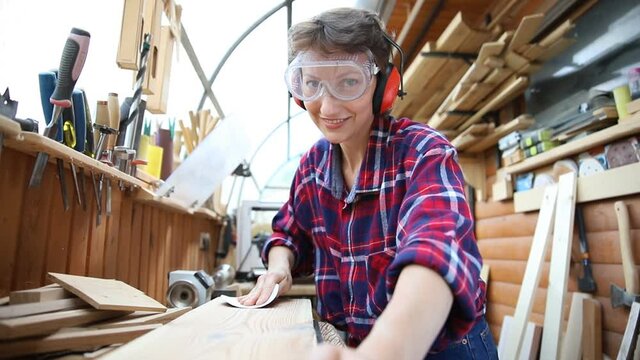 Young attractive 30-40 professional female carpenter looking grinding raw wood with sandpaper in carpentry diy workshop. Feminine women equality concept. Adult girl with male hobby at workbench
