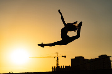Female gymnast jumping showing her flexibility and split during sunset on orange sky background with crane and construction. Concept of freedom and happiness
