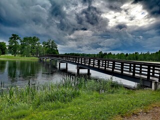 Fototapeta na wymiar Wooden bridge in the park, across a small river with green grass and a dramatic blue sky with dark rain clouds. Summer landscape